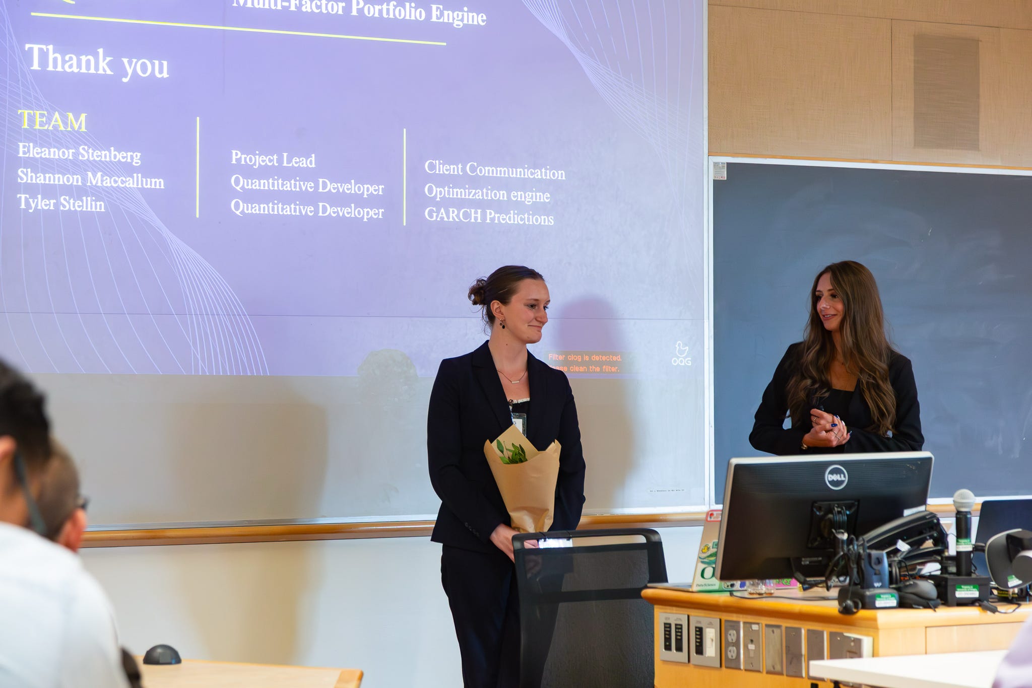 Two members at the front of the room after presenting, with the project team roles on screen
