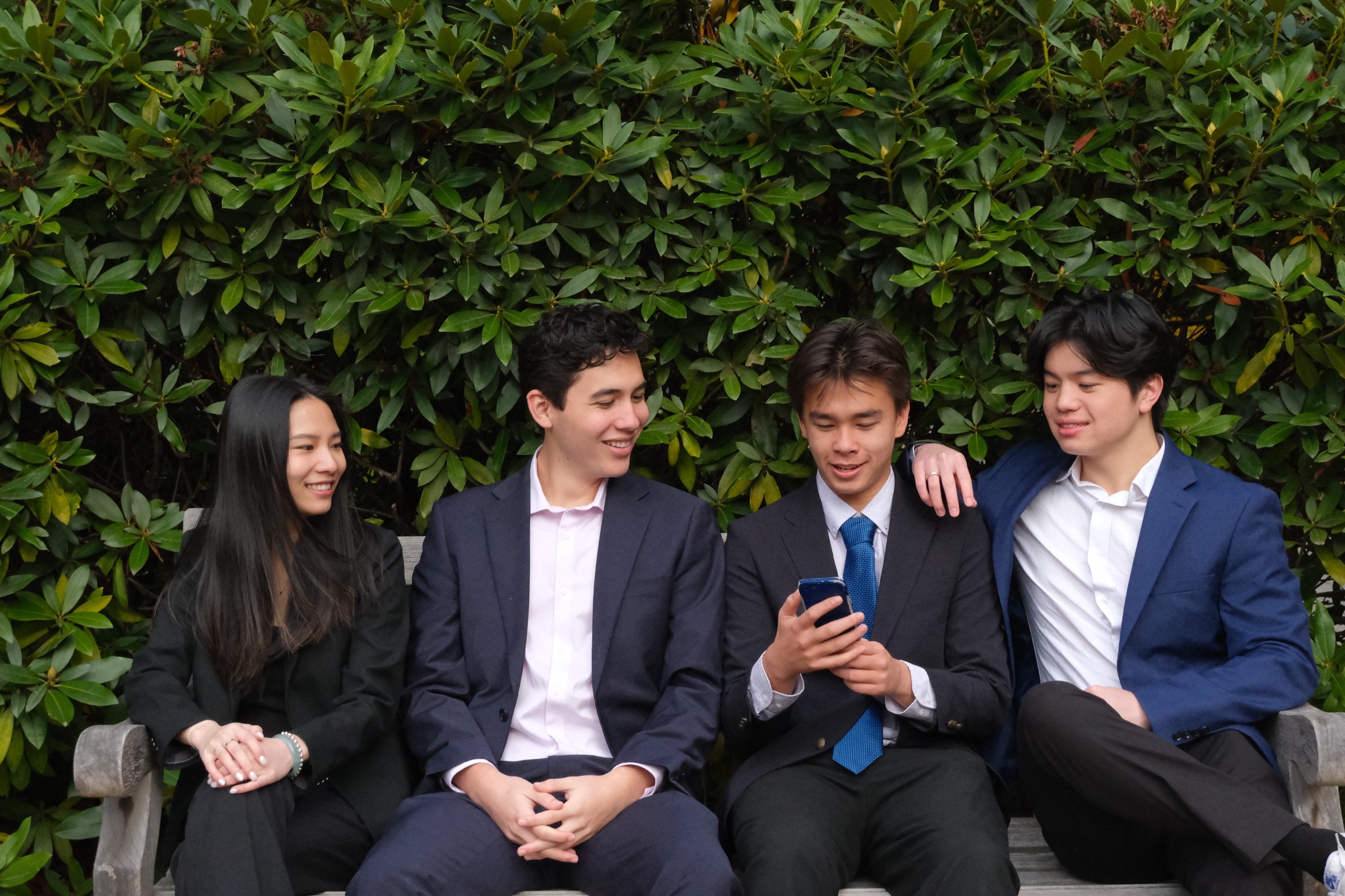 Four members in suits on a bench in front of a hedge